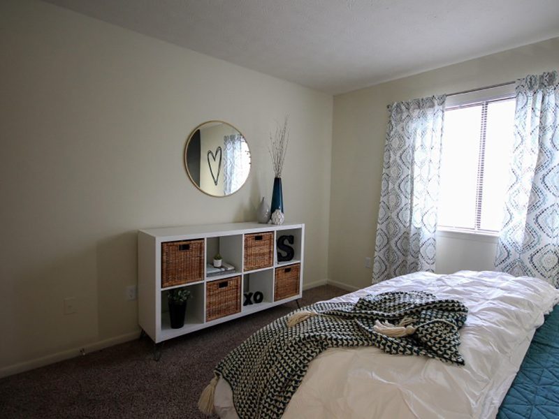 This image shows the bedroom area showcasing its minimal wall decor, comfortable beddings, and a window overlooking the view of TGM Shadeland Station Apartments.